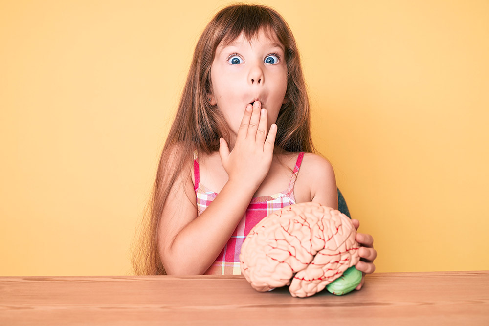 Little caucasian kid girl with long hair holding brain as mental health concept covering mouth with hand, shocked and afraid for mistake. surprised expression